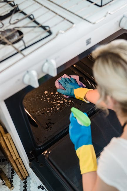 A close-up view of a black oven interior, where a person wearing blue rubber gloves and a yellow sleeve is wiping the oven surface with a purple microfiber cloth. The surface appears to be made of sta