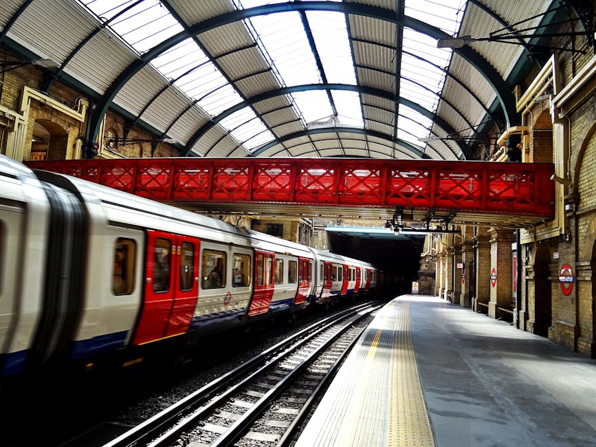 Inside a London underground station with a modern train arriving at the platform. The station features high arched ceilings constructed from metal and glass panels, allowing natural light to illuminat